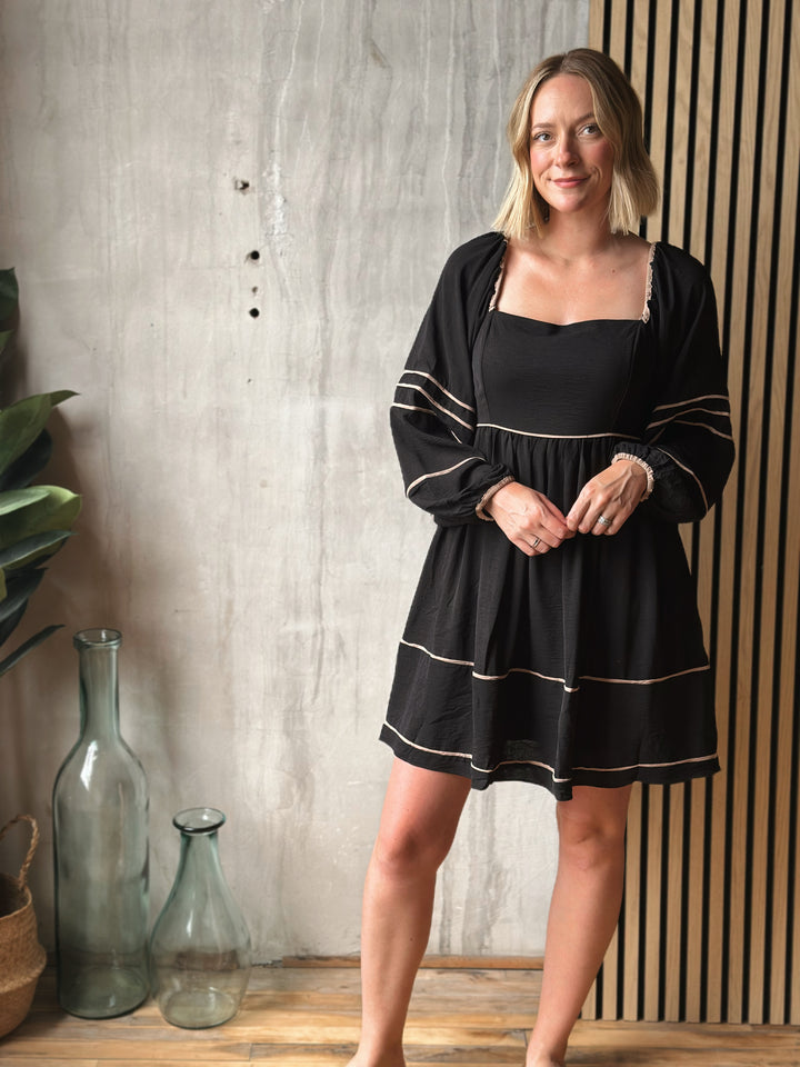 Woman wearing a black dress with white trim standing in a room with decorative vases and plants.