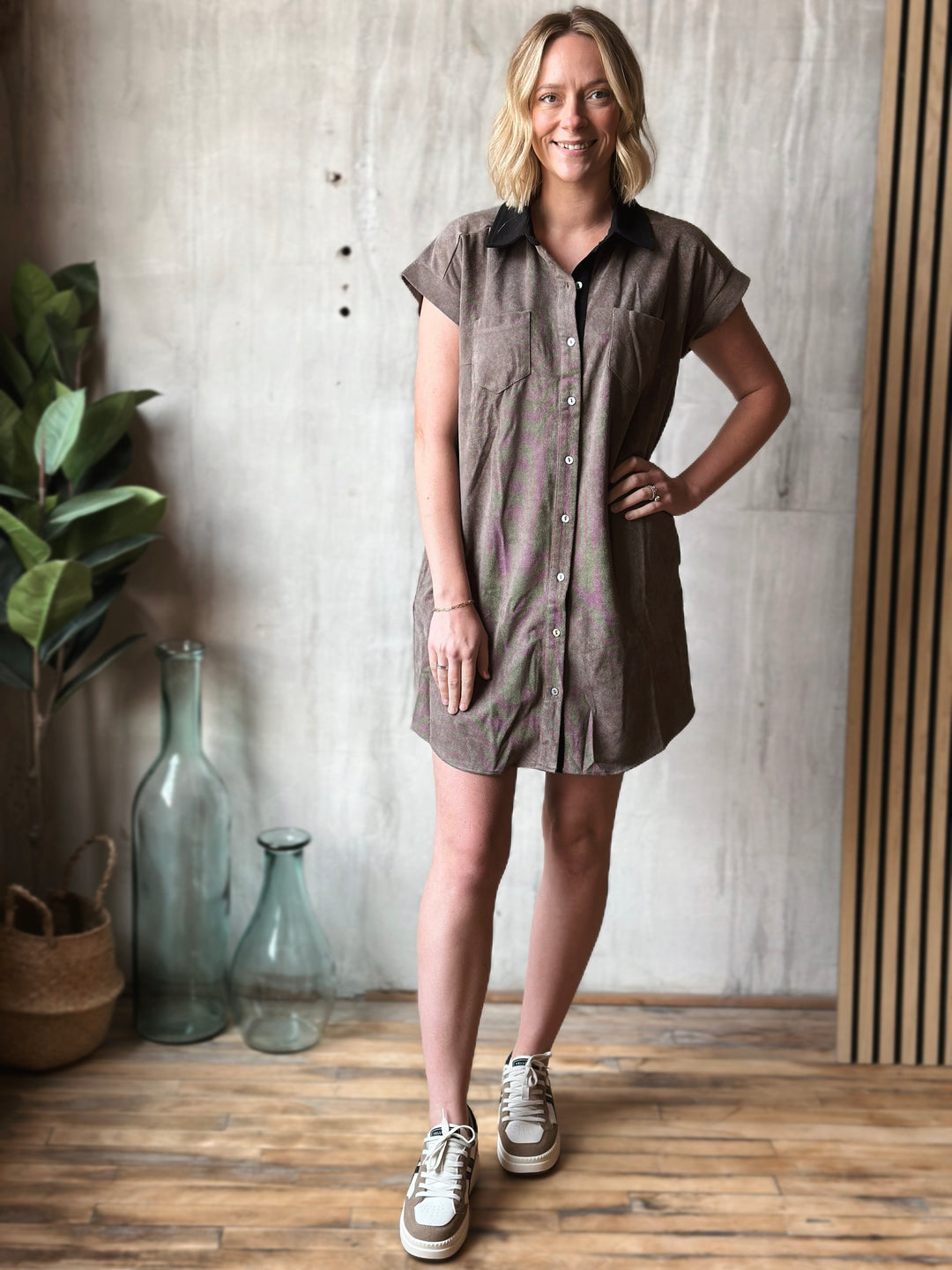 Woman wearing a brown dress standing in a room with a plant and decorative bottles.