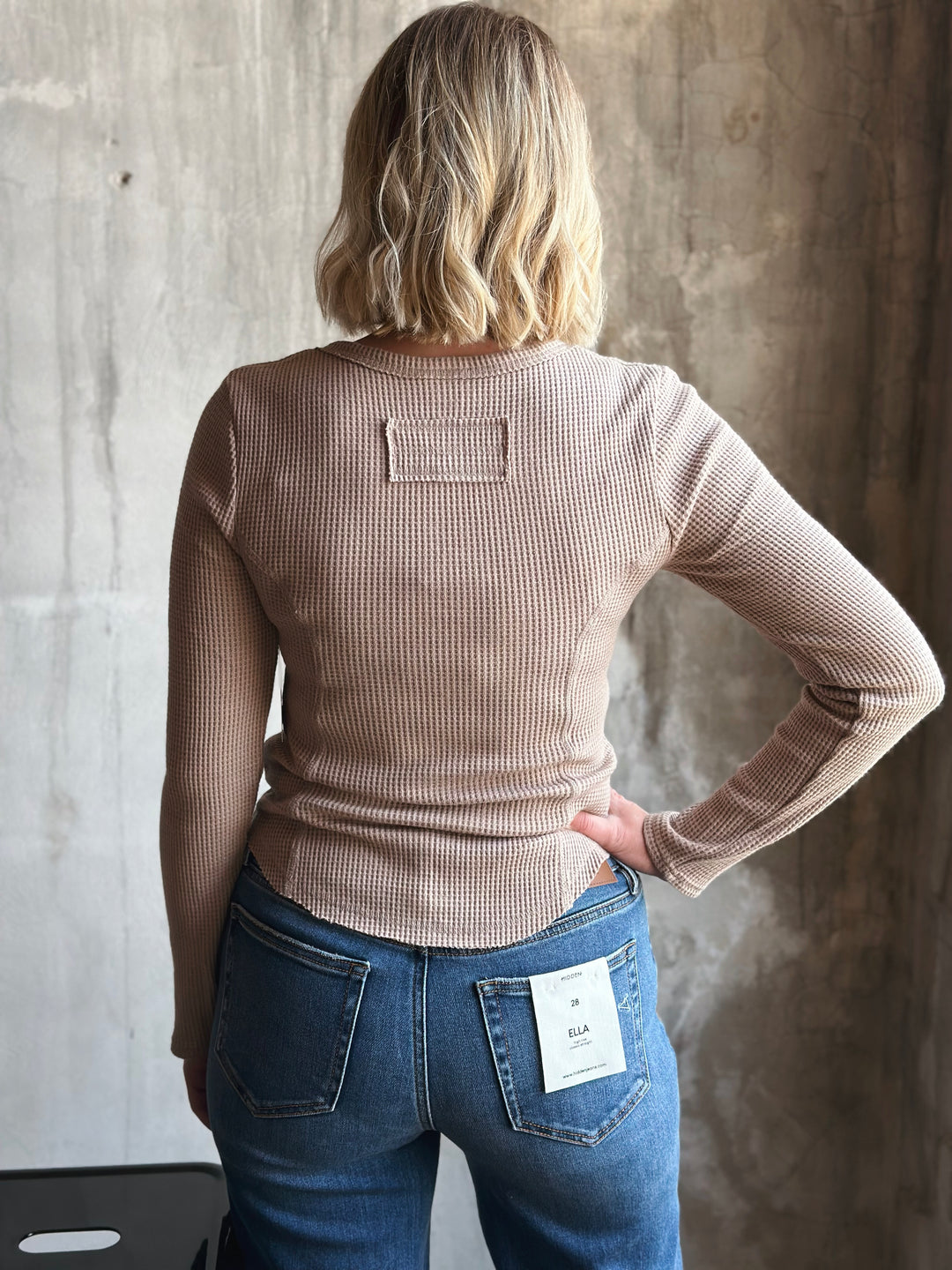 Person wearing a beige ribbed sweater and blue jeans against a textured wall.