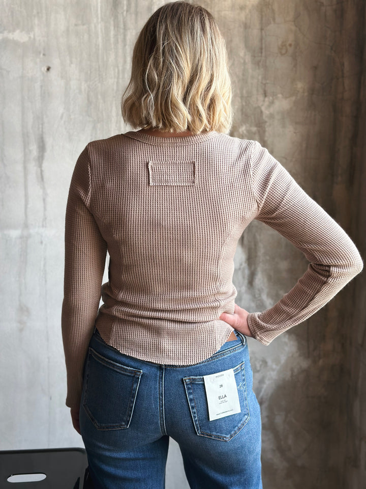 Person wearing a beige ribbed sweater and blue jeans against a textured wall.