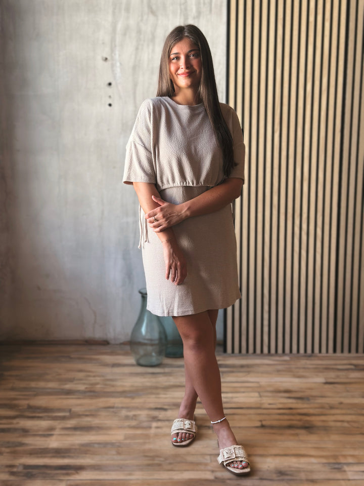 Woman wearing a beige dress standing in a room with wooden flooring and a textured wall.