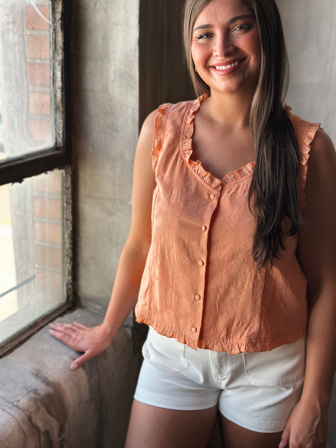 Woman wearing a peach sleeveless top and white shorts standing by a window.