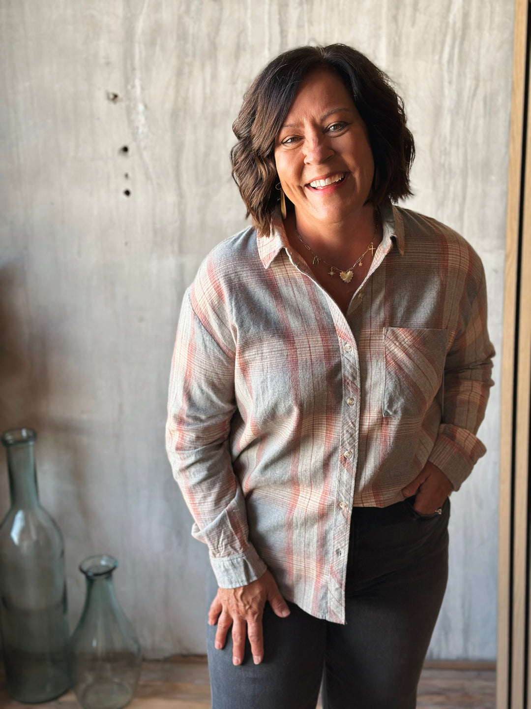 Woman wearing a plaid shirt standing in front of a textured wall with decorative bottles.