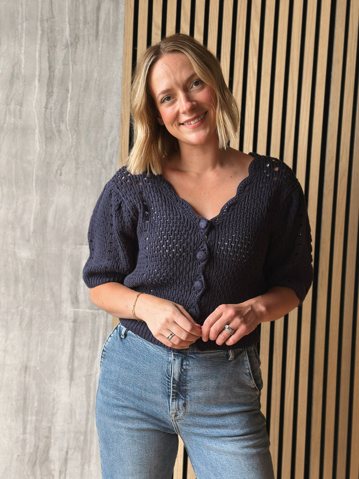 Woman wearing a dark blue knitted cardigan and light blue jeans standing against a wooden panel background.