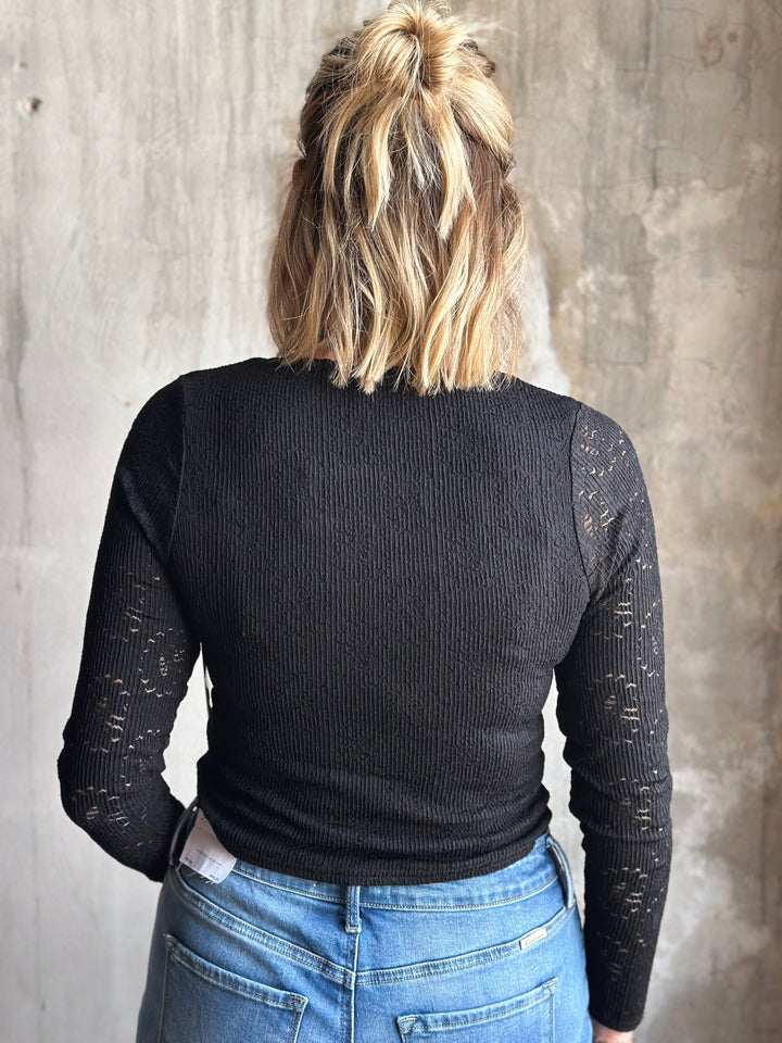 Person wearing a black ribbed sweater with sheer sleeves against a gray concrete wall.