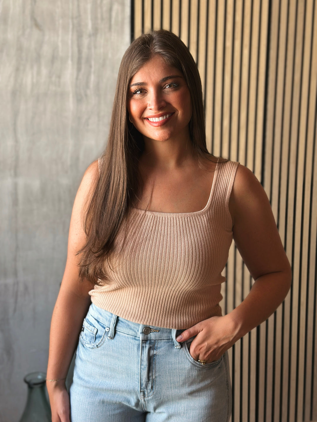 Woman wearing a beige ribbed tank top and light blue jeans against a wooden paneled wall.