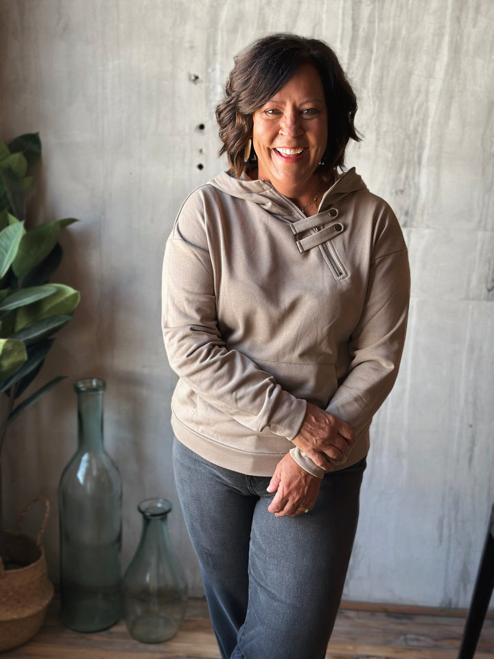 Woman wearing a beige hoodie standing in a room with decorative vases and plants.