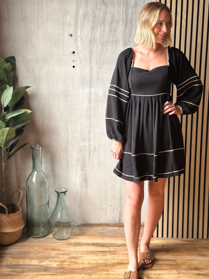 Woman wearing a black dress with white trim standing in a room with a plant and decorative vases.
