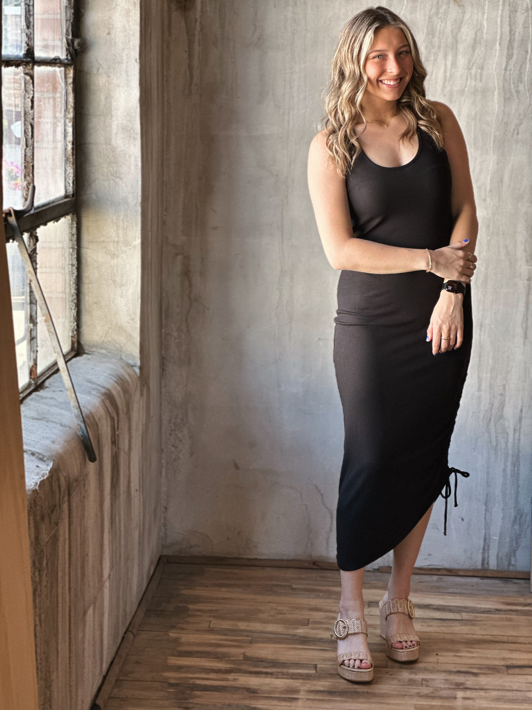 Front view of model wearing a black midi dress with a scoop neckline, sleeveless design and adjustable drawstrings along with a knotted open back detail