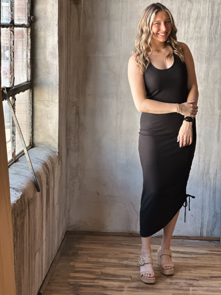 Front view of model wearing a black midi dress with a scoop neckline, sleeveless design and adjustable drawstrings along with a knotted open back detail