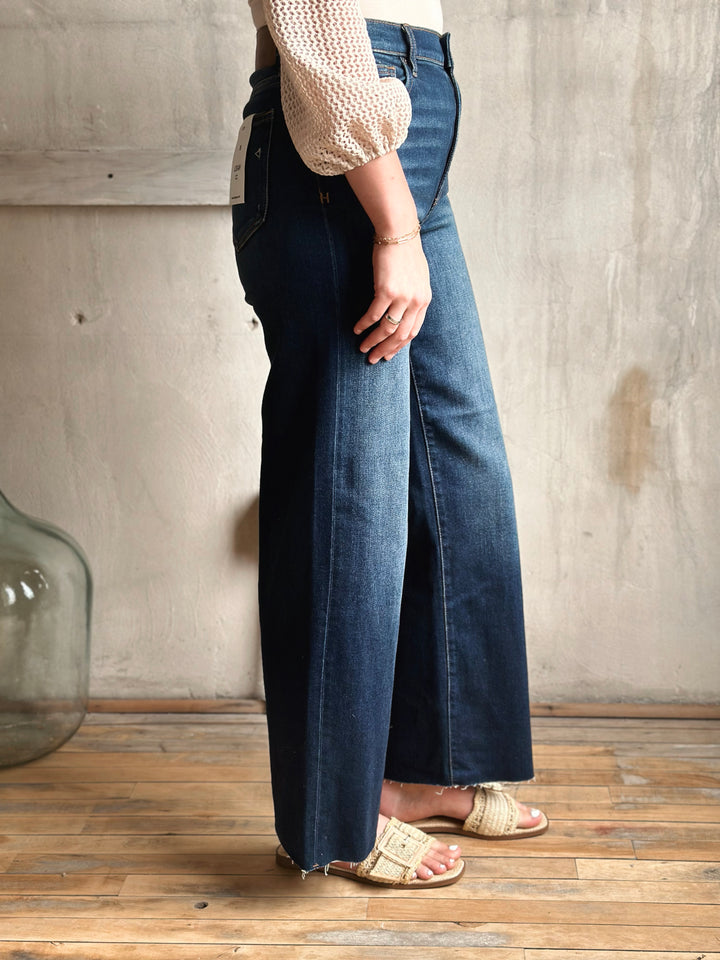 Person wearing blue jeans and a white top against a wooden floor and wall background