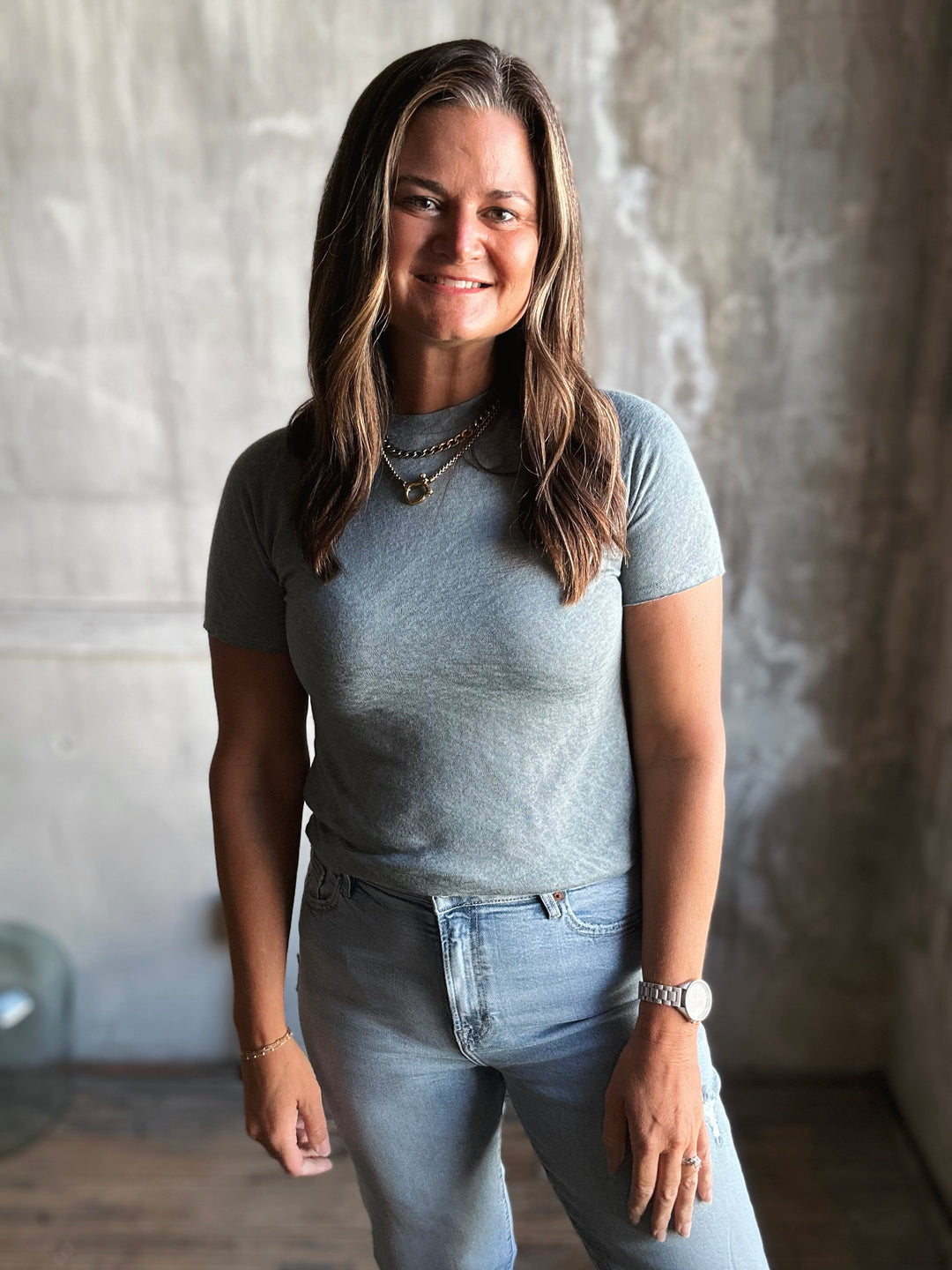 Woman wearing a gray t-shirt and blue jeans standing against a textured wall.