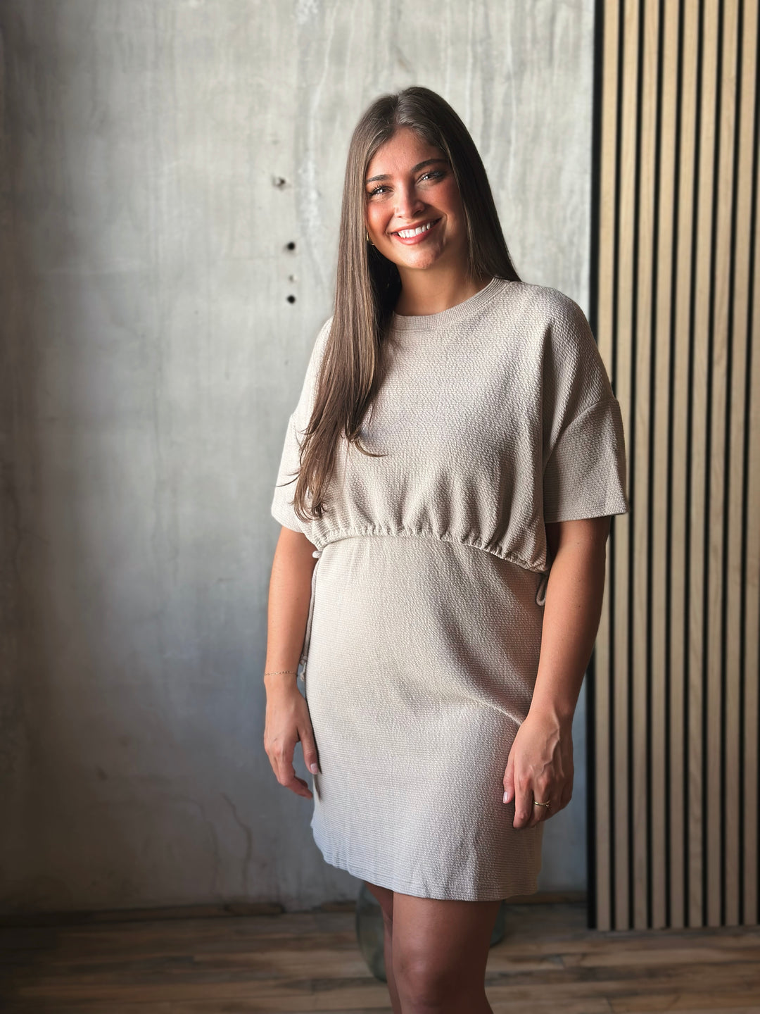 Woman wearing a beige dress standing against a textured wall.