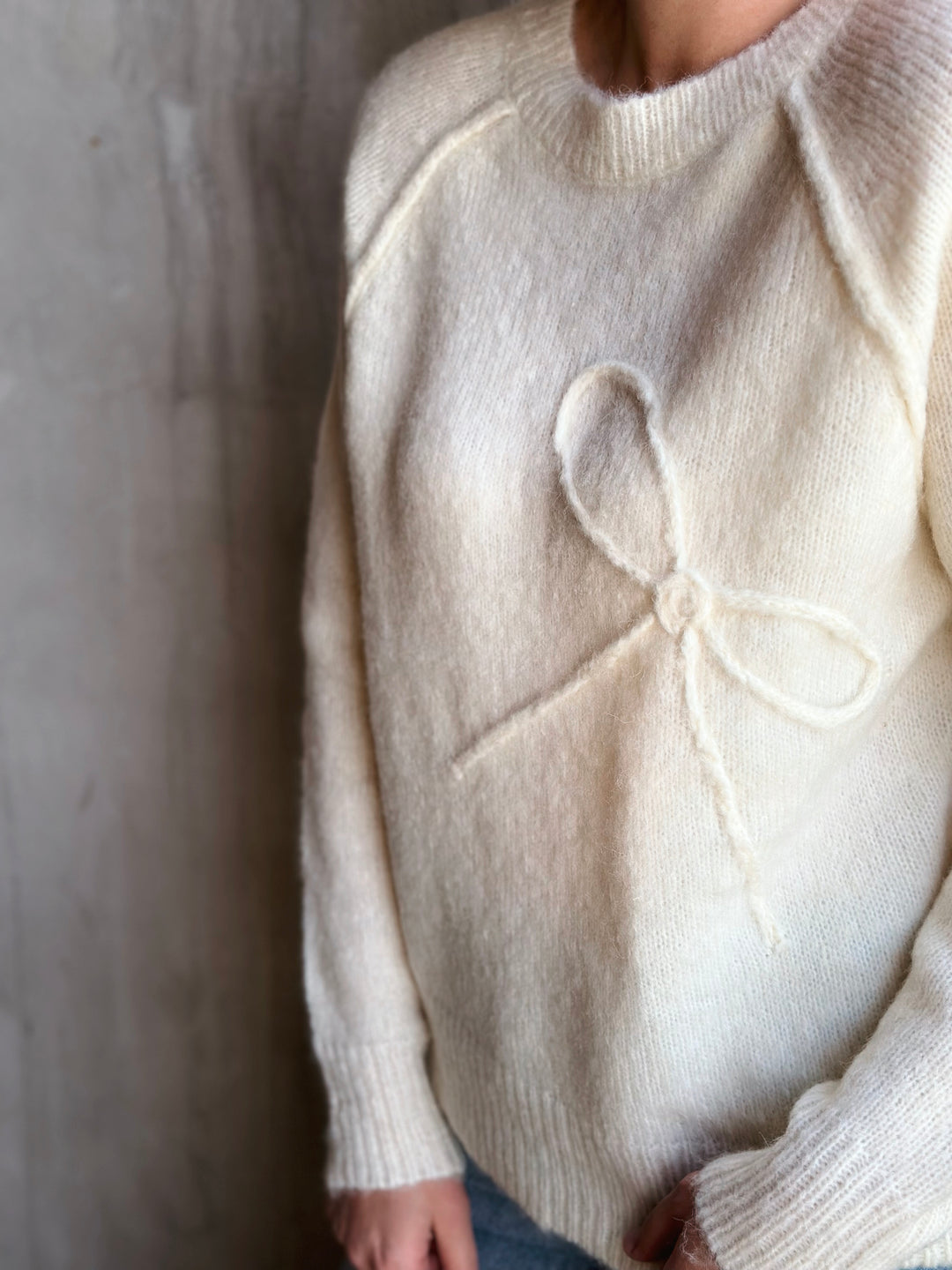 White sweater with a bow design worn by a person against a neutral background
