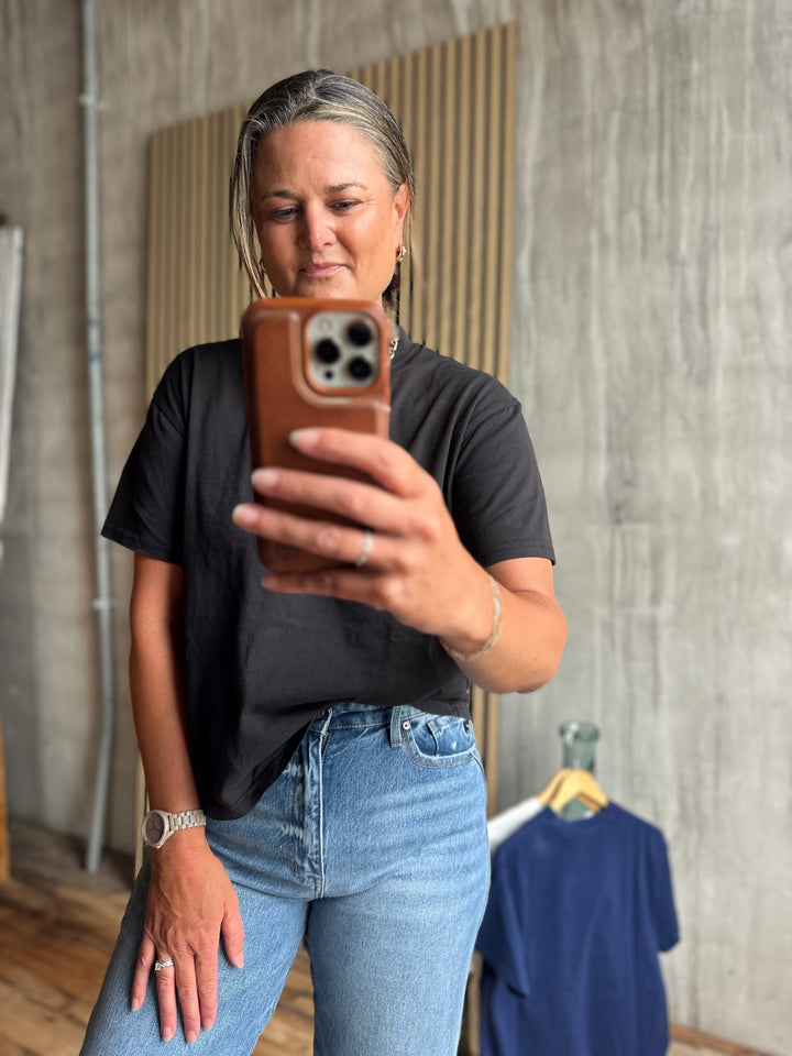 Person taking a mirror selfie wearing a black t-shirt and blue jeans, holding a phone with a brown case.