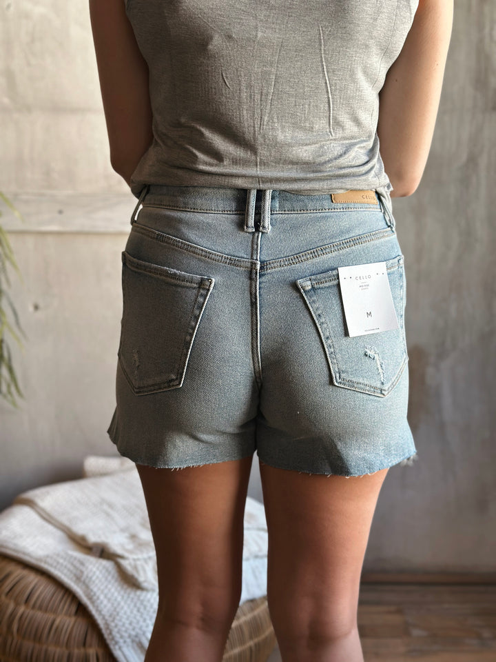 Back view of model wearing mid rise light wash denim shorts that are a midi length with a side slit, there is also pockets on front and back with a button and fly closure
