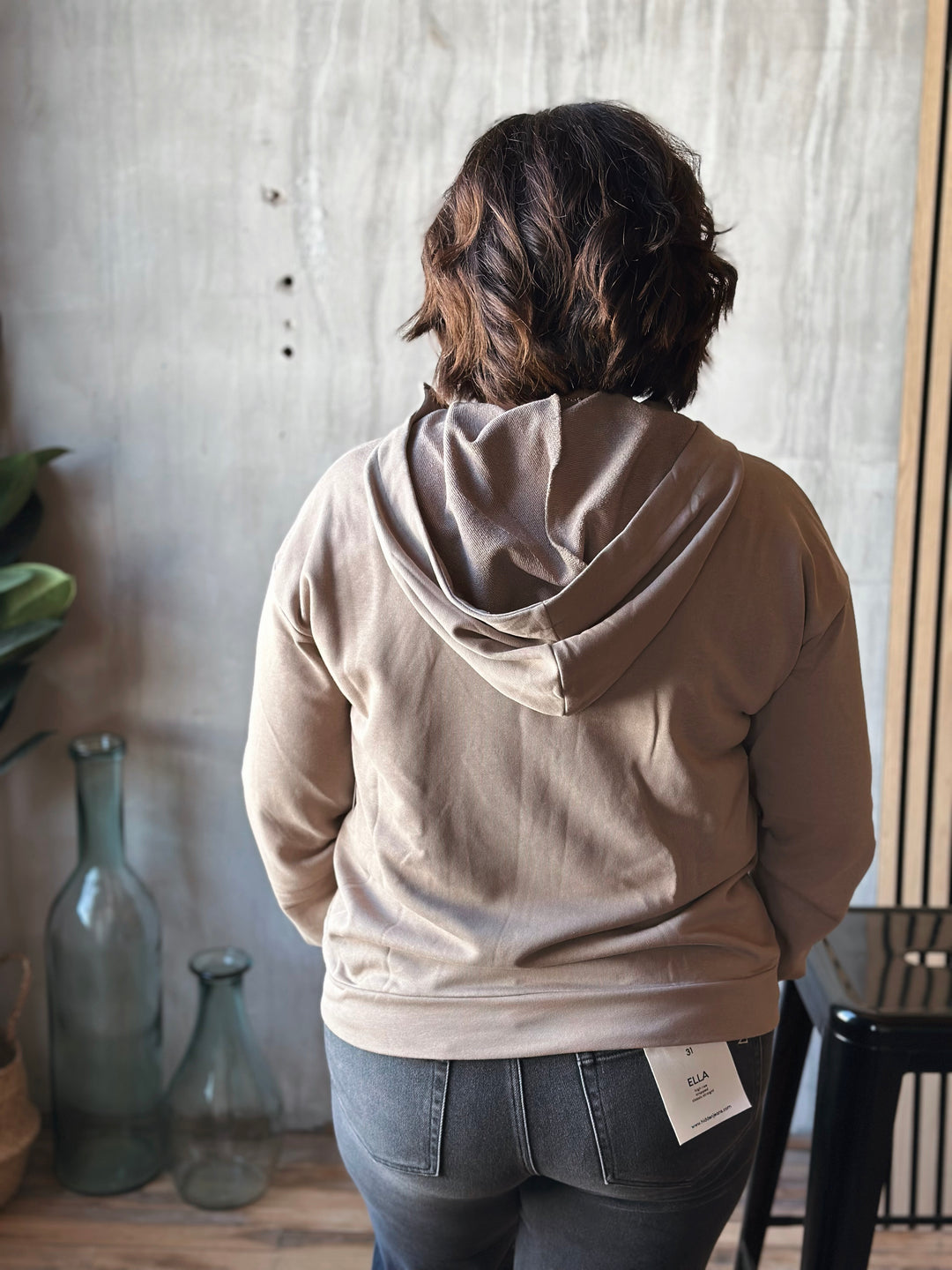 Person wearing a beige hoodie with a tag, standing in a room with a textured wall and decorative bottles.