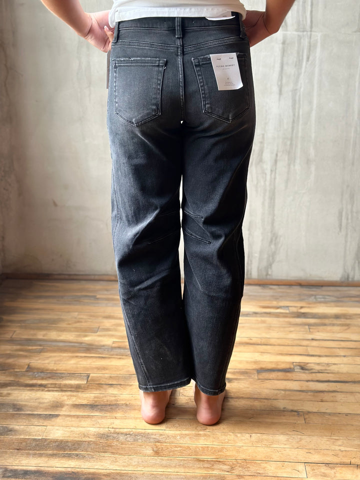 Rear view of a person wearing dark gray washed denim jeans with a white paper tag still attached to the back pocket, standing barefoot on a wooden floor against a concrete wall.