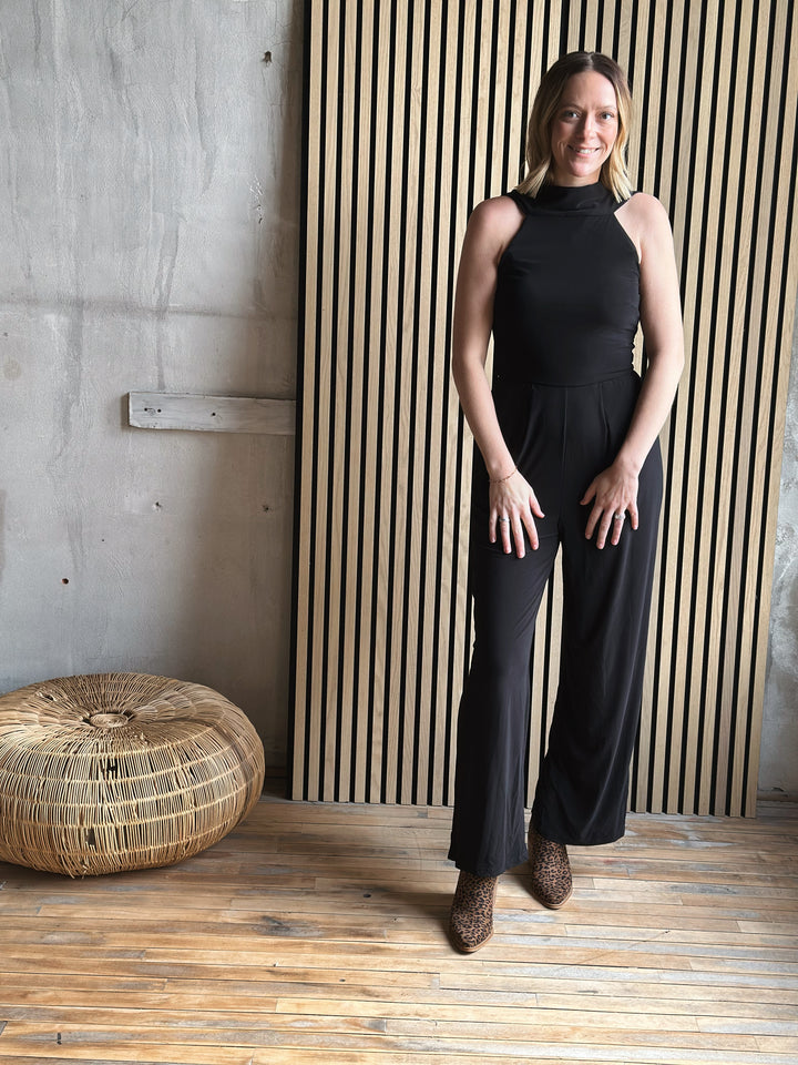 Woman in a black jumpsuit standing in a room with wooden flooring and a textured wall.