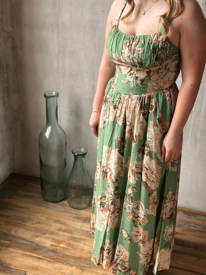 Woman wearing a green floral dress standing in a room with a textured wall and wooden floor.