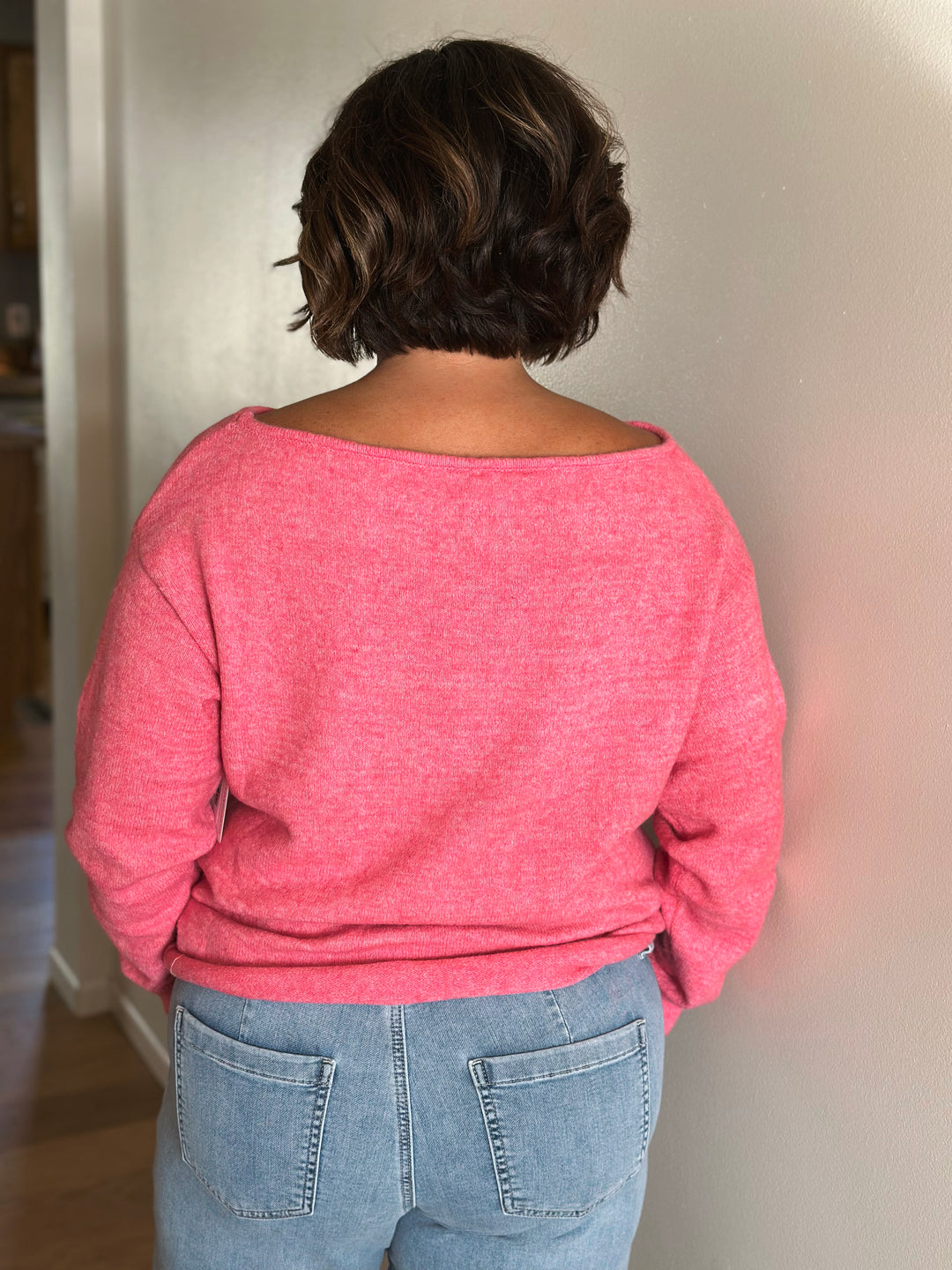 Person wearing a pink sweater and blue jeans standing against a plain wall.