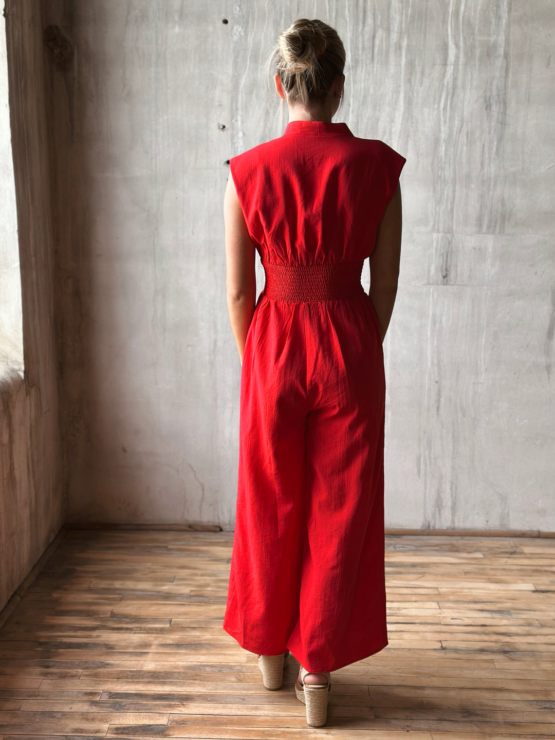 Back view of sleeveless red jumpsuit features a flattering v-neckline, a front zipper closure, and a smocked waist