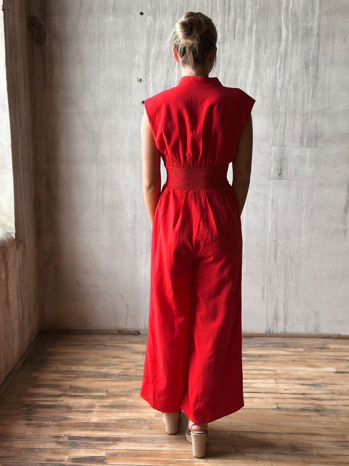 Back view of sleeveless red jumpsuit features a flattering v-neckline, a front zipper closure, and a smocked waist