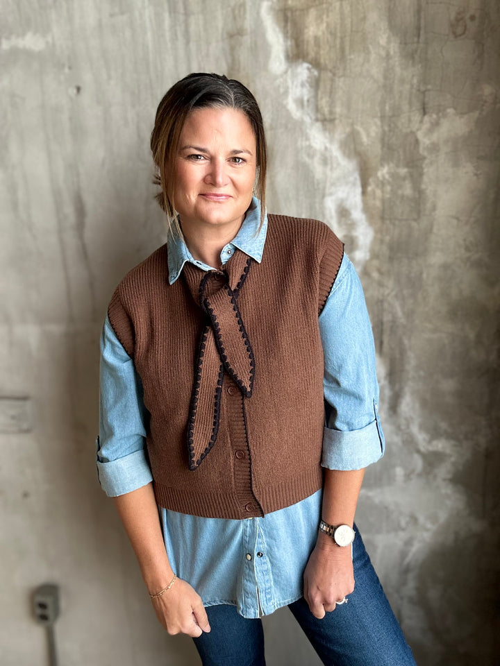 A woman wearing a brown knit button-front vest with a tie-neck detail over a light blue denim shirt, paired with blue jeans, standing against a textured concrete wall.