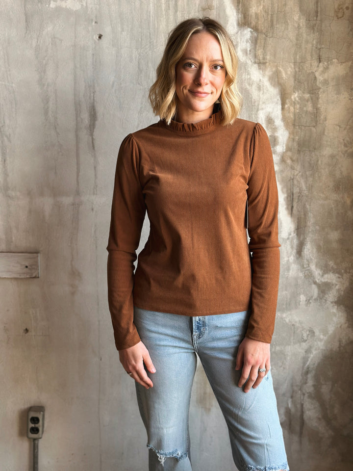 Woman wearing a brown long-sleeve top and light blue jeans against a textured wall.