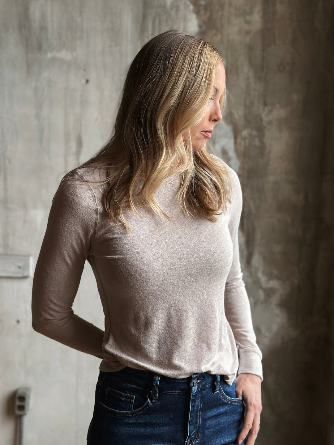 Woman wearing a beige long-sleeve top and blue jeans against a textured wall.