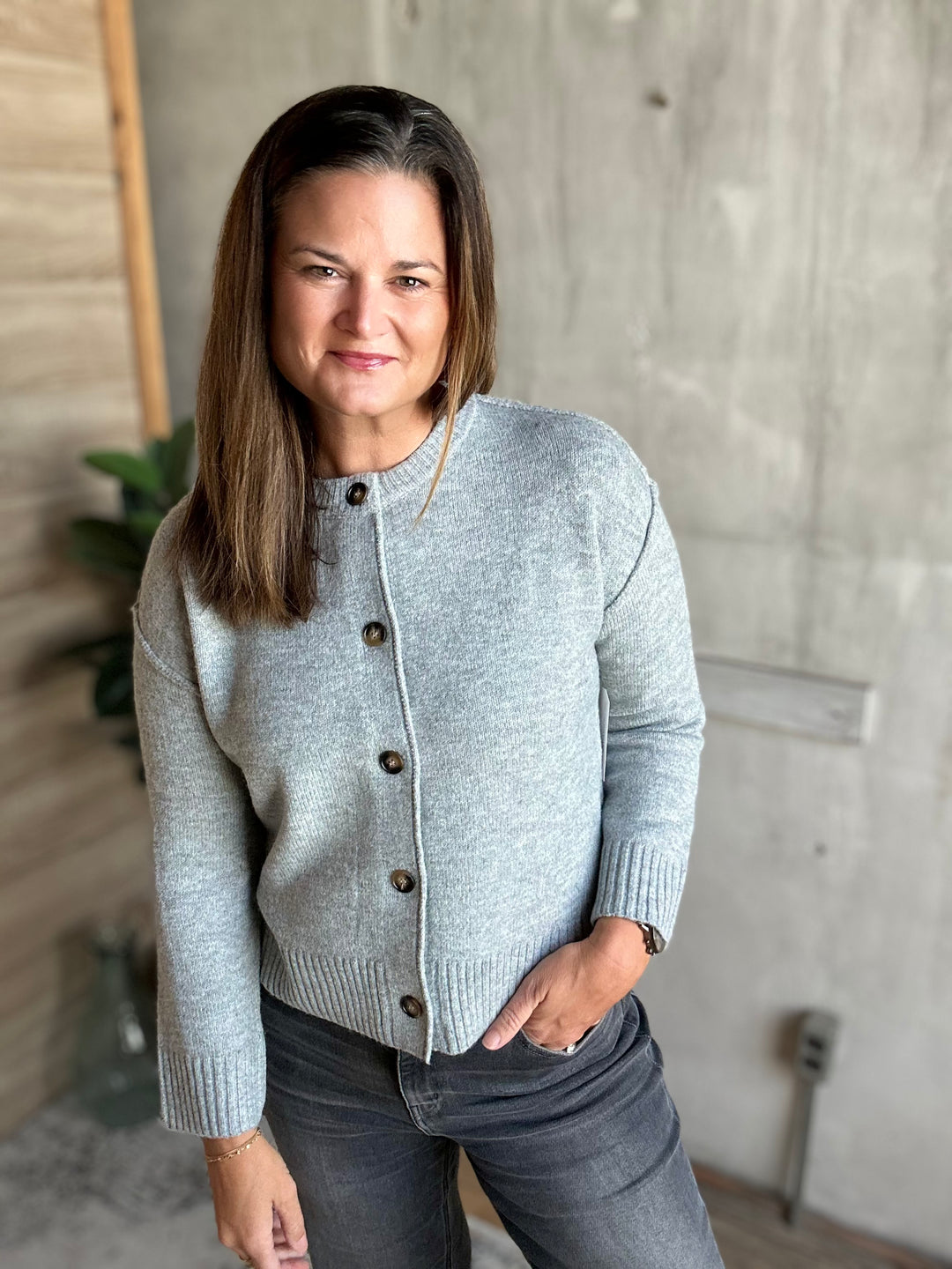 A woman wearing a light gray knit cardigan sweater with dark buttons, paired with dark gray jeans, standing in a modern room with a concrete wall background.
