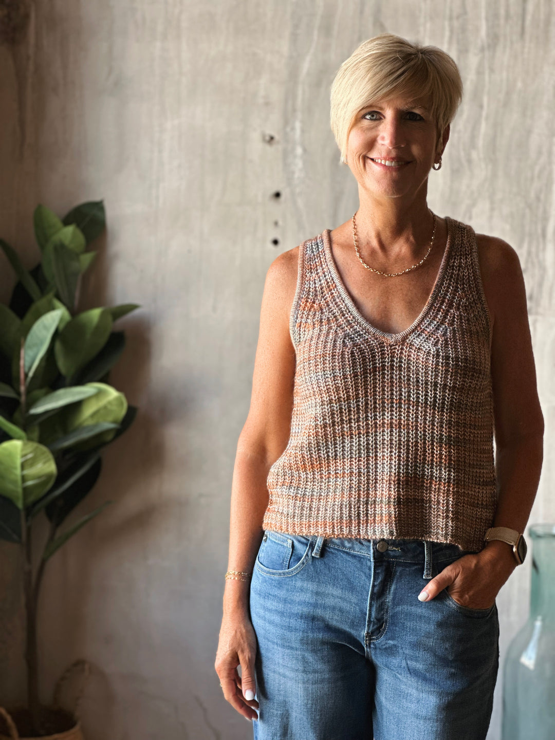 Woman wearing a patterned tank top and blue jeans standing against a concrete wall with a plant to the left.