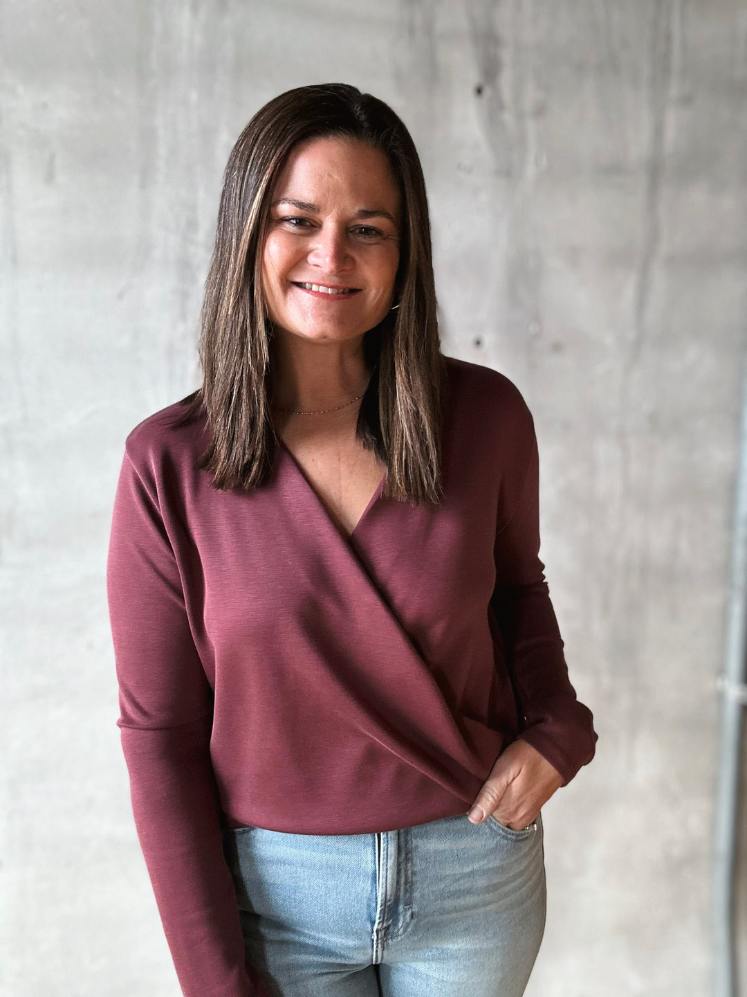 Woman wearing a maroon long-sleeve top and light blue jeans against a gray concrete wall.