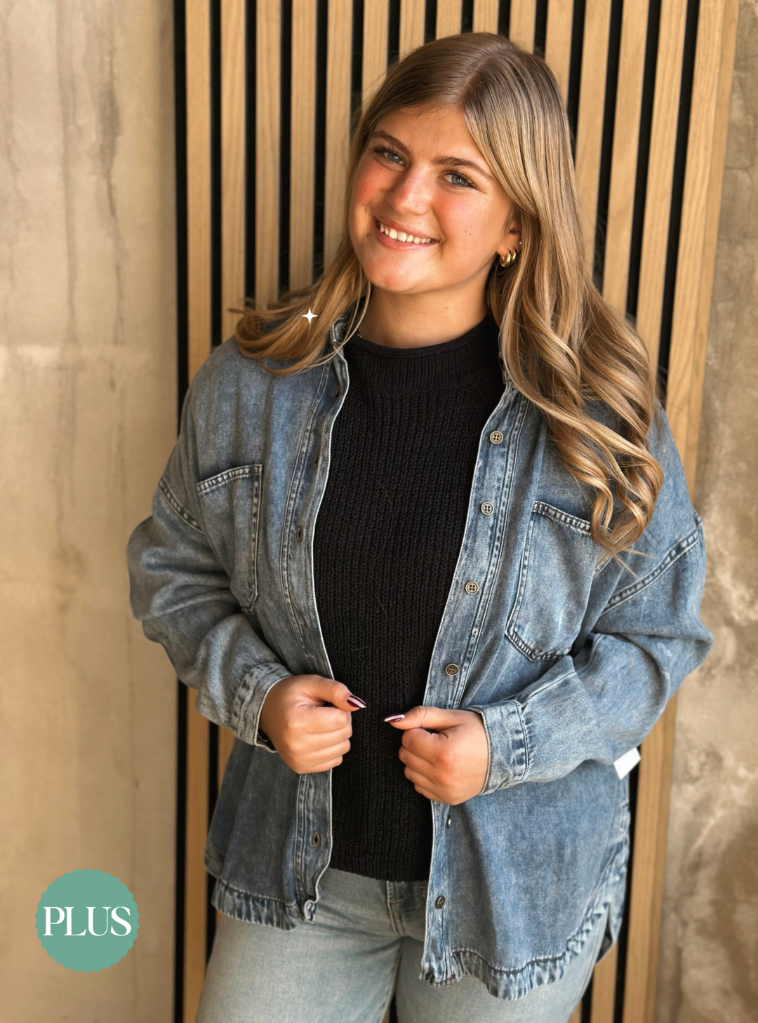 Woman wearing a denim jacket over a black sweater, standing against a wooden panel background.