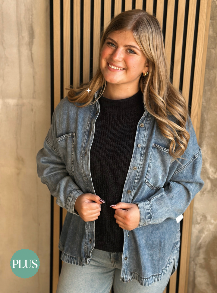 Woman wearing a denim jacket over a black sweater, standing against a wooden panel background.