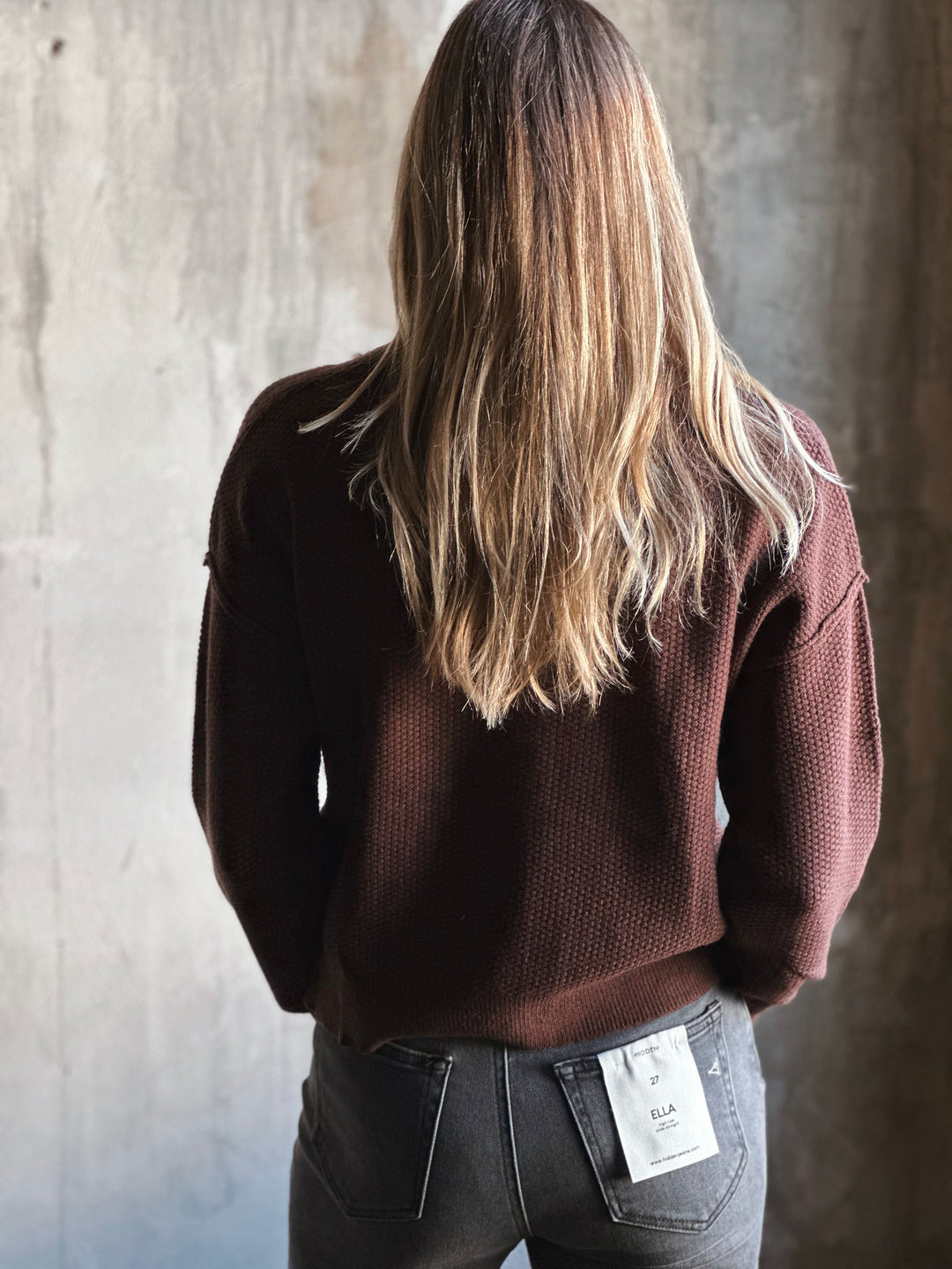 Person wearing a brown sweater and gray jeans against a textured wall.
