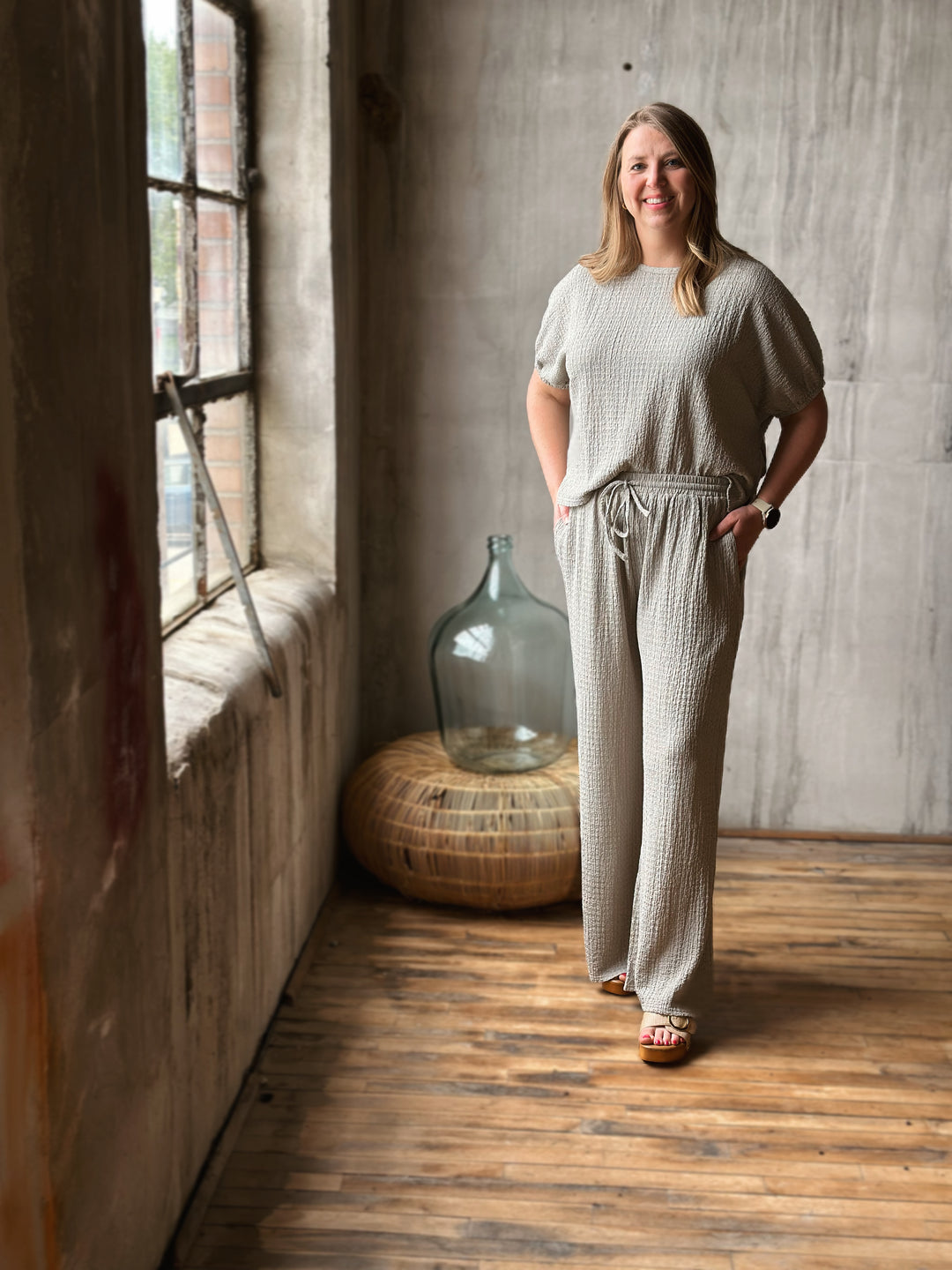 Woman in a textured sage outfit standing in a room with wooden floor and large window.