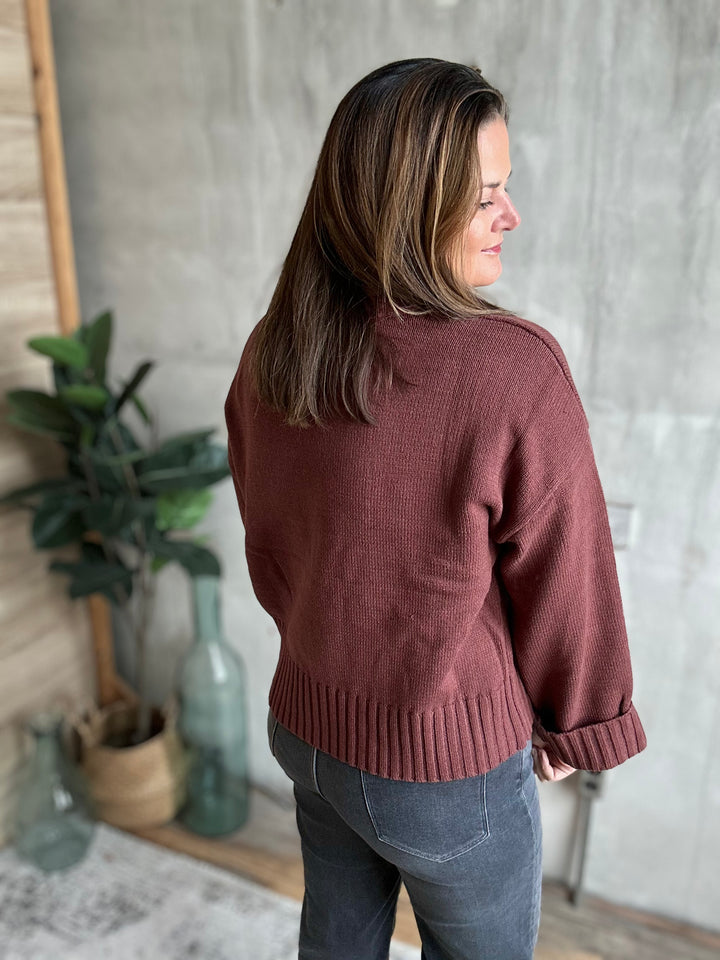 A woman with shoulder-length brown hair is shown from the back wearing a dusty rose ribbed knit sweater and dark gray jeans, standing in a room with a concrete-textured wall, wooden paneling, and potted plants.