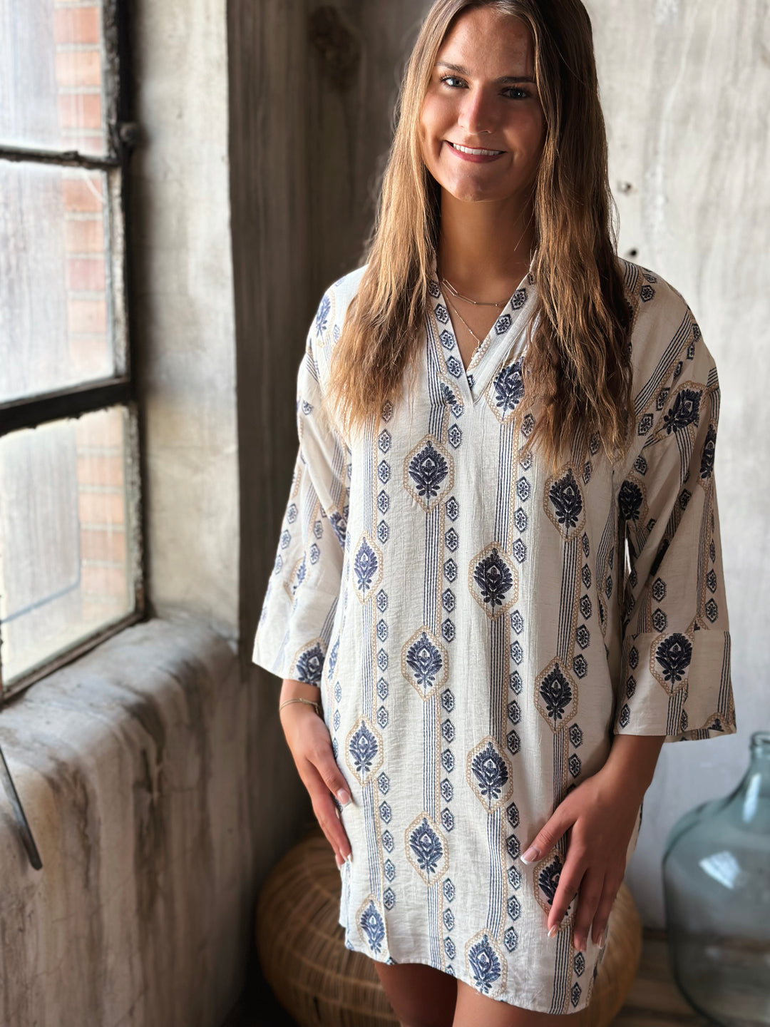 Woman wearing a white patterned v-neck dress standing in a room with a large window.