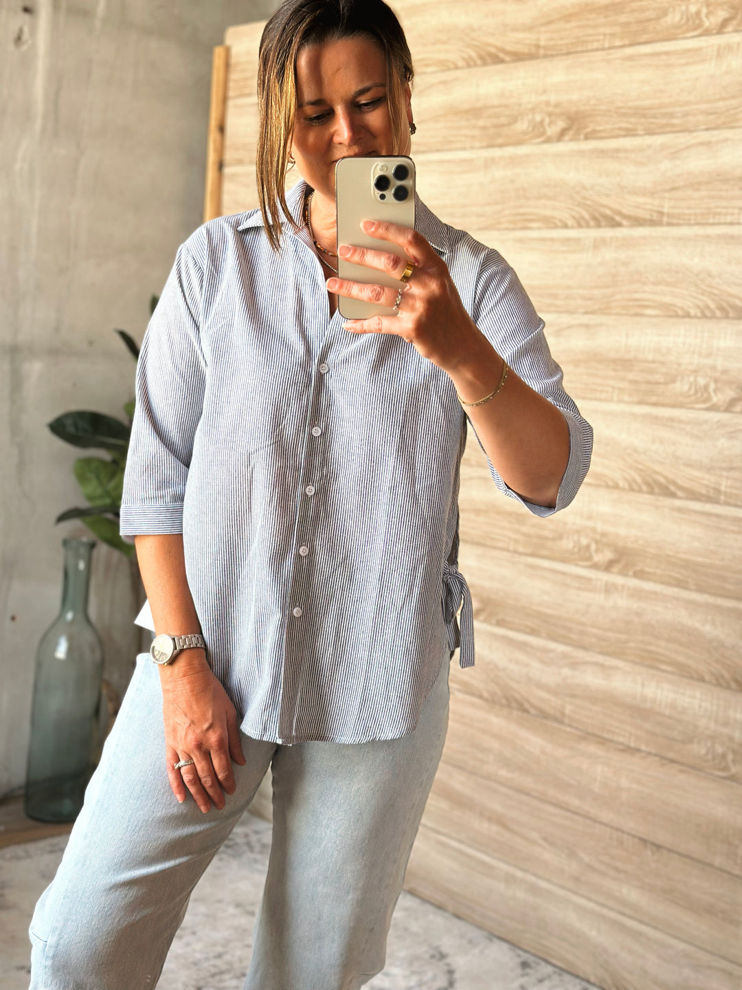Person taking a mirror selfie wearing a light gray shirt and pants in a room with a wooden wall.