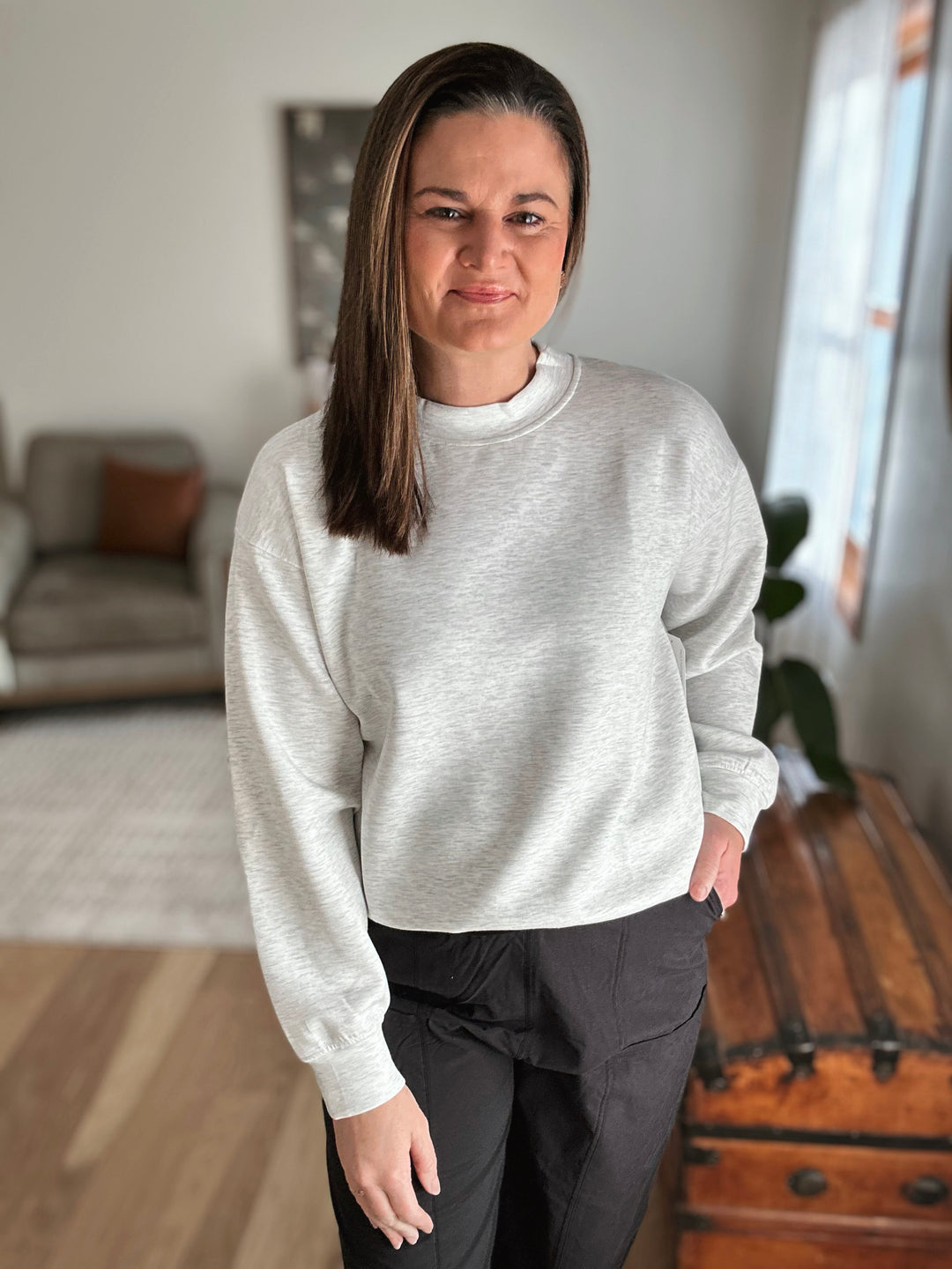Person wearing a gray sweater and black pants standing in a room with a couch and window in the background.