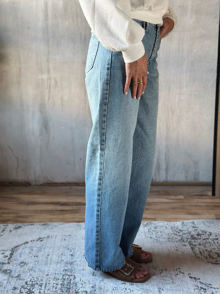 Person wearing light blue jeans and a white shirt in a room with a textured wall and wooden floor.