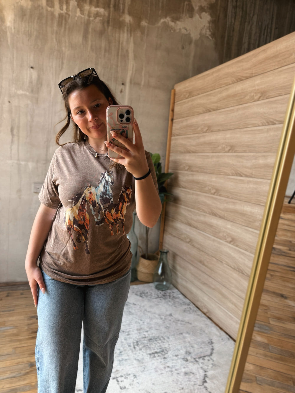 A person taking a mirror selfie wearing a graphic t-shirt with three horses on it in a room with wooden flooring and a concrete wall.