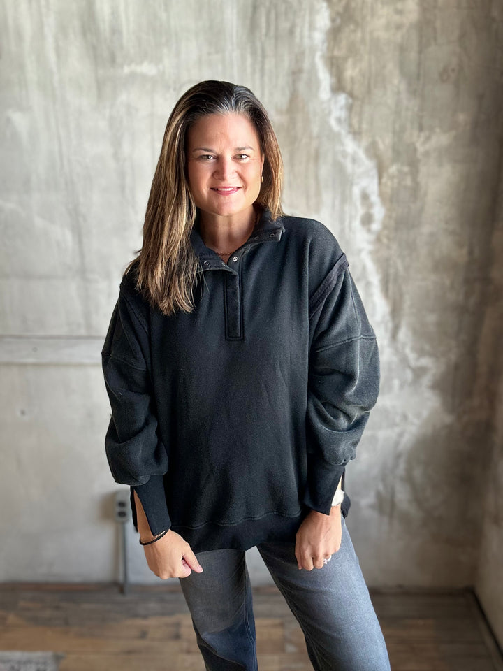 A woman with shoulder-length brown hair stands indoors wearing a dark charcoal gray half-placket pullover sweatshirt and gray jeans, posing in front of a textured concrete wall.
