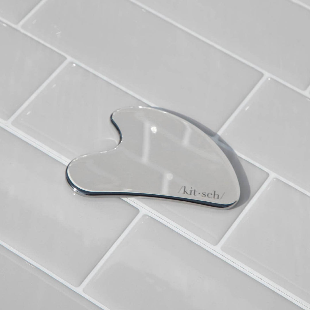 Glass gua sha tool with 'kitsch' branding on a tiled bathroom floor.