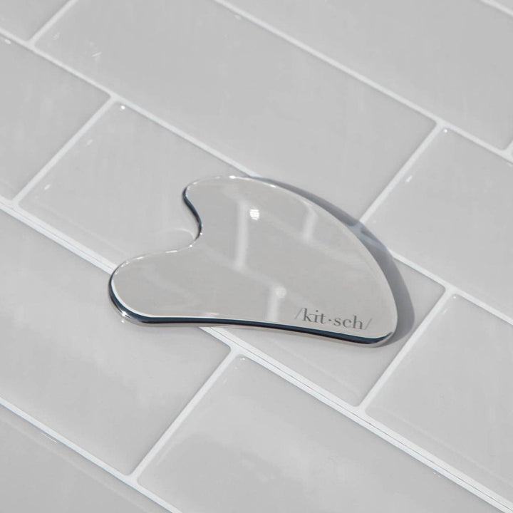 Glass gua sha tool with 'kitsch' branding on a tiled bathroom floor.