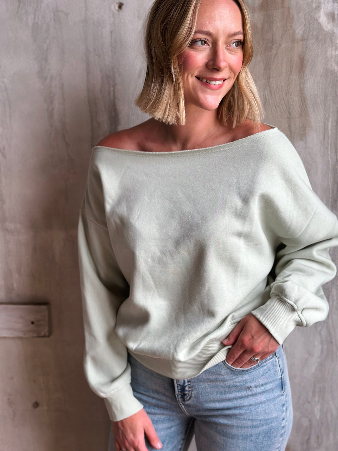 Woman wearing a light-colored off-shoulder sweater and jeans against a textured wall.