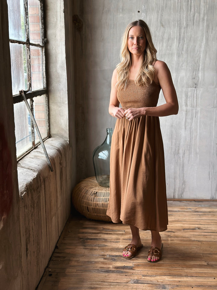 Woman in a brown dress standing in a room with wooden floor and large window.