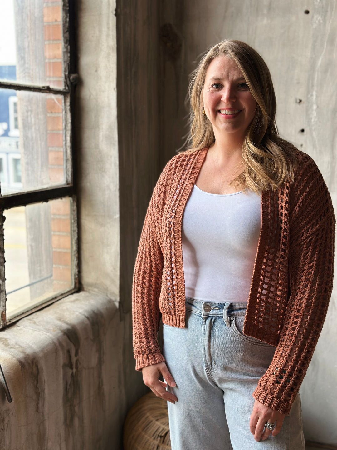 Woman wearing a light brown knitted cardigan standing by a window with a concrete wall background.