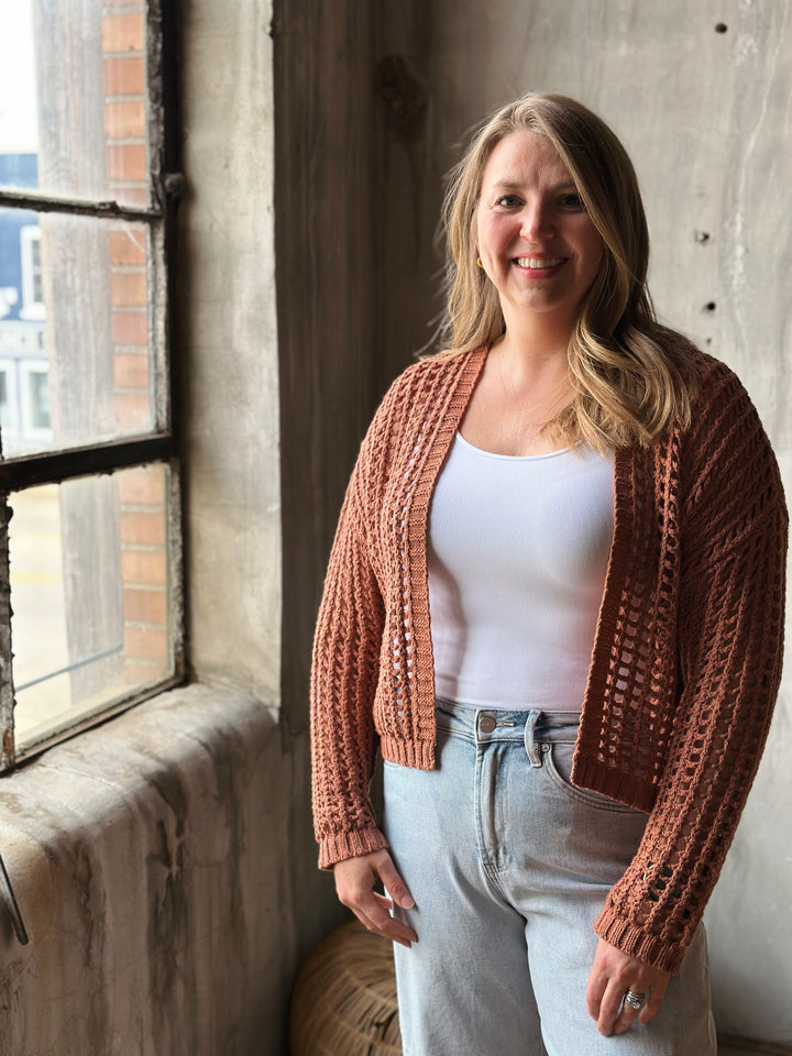 Woman wearing a light brown knitted cardigan standing by a window with a concrete wall background.